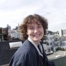 Smiling young woman with curly hair sitting on a rooftop overlooking a cityscape.